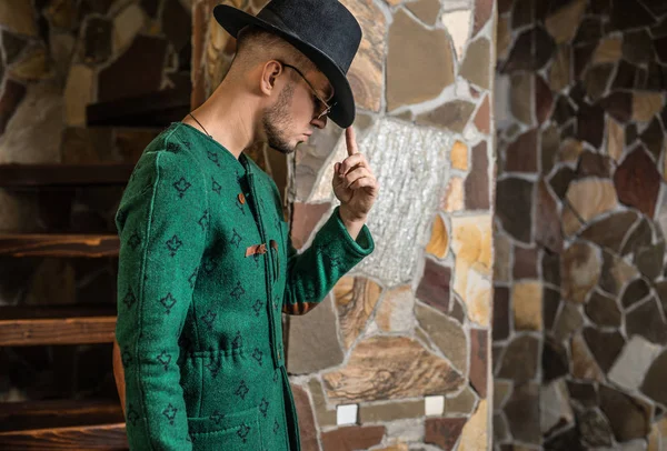 Guapo joven hombre con estilo en abrigo verde largo y sombrero sobre fondo de piedra . — Foto de Stock