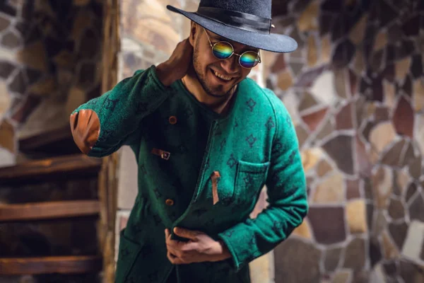 Handsome young stylish man in long green coat and hat against stone background. — Stock Photo, Image