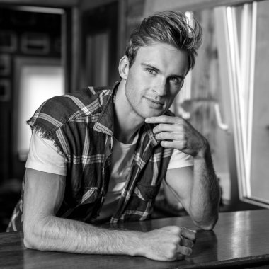 Rahat kıyafetler içindeki yakışıklı genç adam evin içinde poz veriyor. Siyah-beyaz portre.