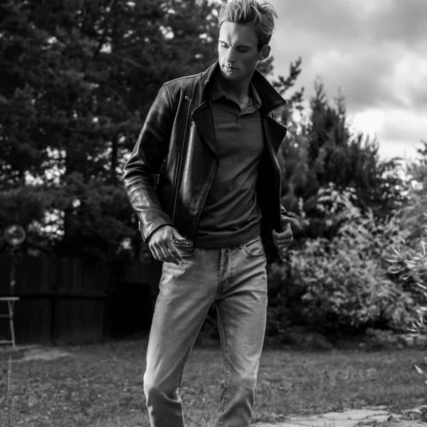Ein junger gutaussehender Mann in klassischer Lederjacke posiert im Sommergarten. Schwarz-Weiß-Foto. — Stockfoto