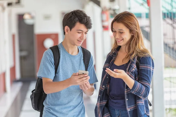 Подростковая пара улыбается, глядя на смартфон в школьном коридоре — стоковое фото
