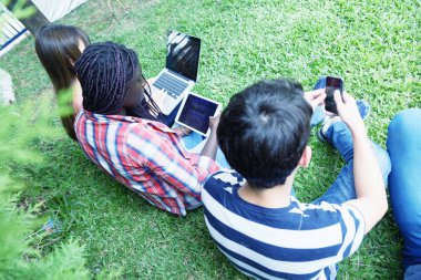Çok etnik gençler arkadaş bir parkın oturmuş elektronik araçları kullanma.