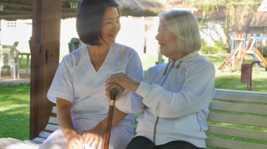 Asyalı doktor, yaşlı ve emekli bir kadınla konuşuyor. İkisi de bahçe bankında oturuyor. Mutluluk ve emeklilik kavramı.