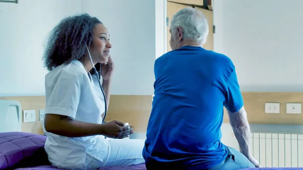 非洲女医生帮助住在医院病床上的老年男病人 使用听诊器进行健康检查 康复和退休概念 — 图库照片