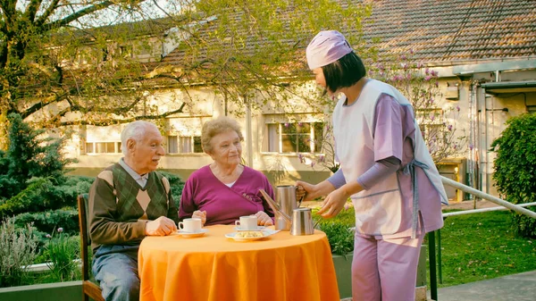 年轻的亚洲女服务员在一家医院的康复花园为年迈的退休夫妇提供早餐 — 图库照片