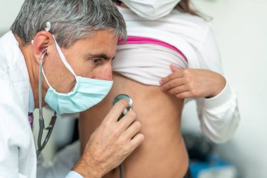 Doktor maske takıyor ve steteskopla Coronavirus döneminde genç bir kızı ziyaret ediyor..