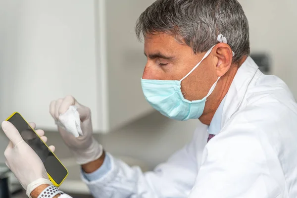 在验尸官时代 疲惫的医生戴着面具和手套在医疗演播室里清洁智能手机 — 图库照片