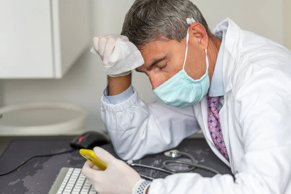 在验尸官时代 戴着面具和手套的疲惫的医生正在医院检查他的智能手机 — 图库照片