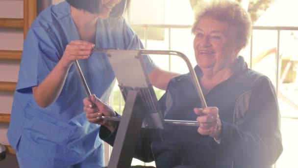 Elder woman making legs exercises at hospital gym with nurse help. — Stock Video