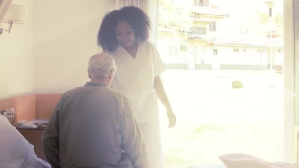 Asyalı hemşire yaşlı bir adamın hastane yatağına gitmesine yardım ediyor. Dışarıdan gün ışığı geliyor. — Stok video