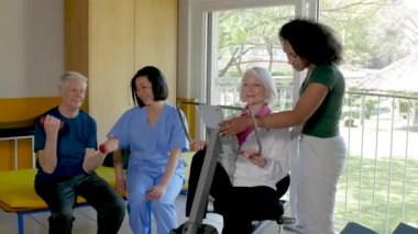 İki genç hemşirenin yardımıyla spor salonunda bacak egzersizi yapan yaşlı çift.