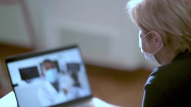 Evde kendini karantinaya almak için maske takan yaşlı bir kadın video görüşmesinde üniforma giyen doktoruyla konuşuyor. PC monitörü bayan omzunun üstünden görünüyor. İnternet üzerinden müşteriye salgın hakkında danışma