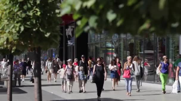 Lidé chodí po ulici. Dav lidí chodit na ulici. Letní slunečný den, lidé na ulici oblečená v letních šatech. V popředí zelené stromy, nikoliv do zaměření. Lidé chodí po ulici. — Stock video