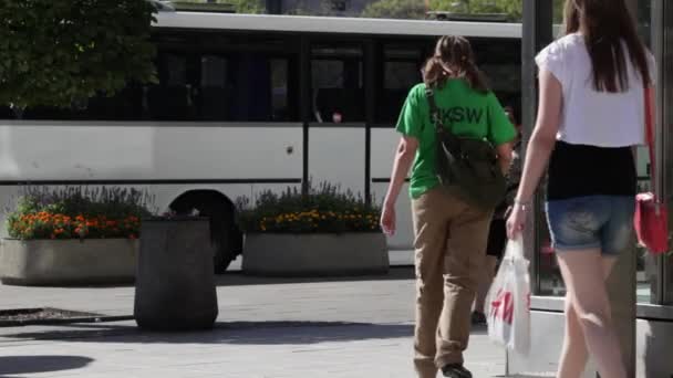 Ludzie chodzą ulicą. Tłum ludzi spaceru na ulicy. Letnim słoneczny dzień, ludzie na ulicy ubrani w letnie ubrania. Na pierwszym planie zielone drzewa, nie w centrum uwagi. Ludzie chodzą ulicą. — Wideo stockowe