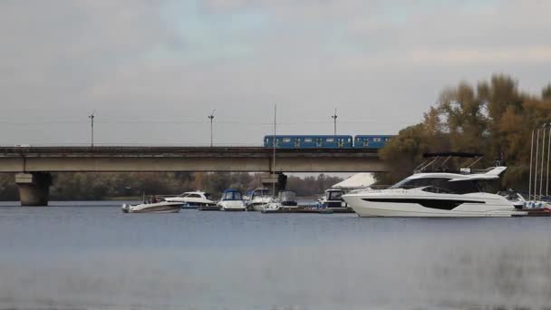 Boats and yachts in the port. Kyiv Ukraine. Bridge over river. White boat on a background of yellow trees and a bridge. Autumn yellow trees. In the background the bridge, passing subway train — Stock Video