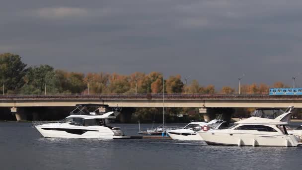 Boats and yachts in the port. Kyiv Ukraine. Bridge over river. White boat on a background of yellow trees and a bridge. Autumn yellow trees. In the background the bridge, passing subway train — Stock Video