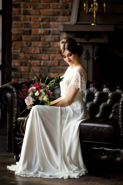 Bride in beautiful dress sitting on sofa indoors in studio interior like at home. — Stock Photo, Image