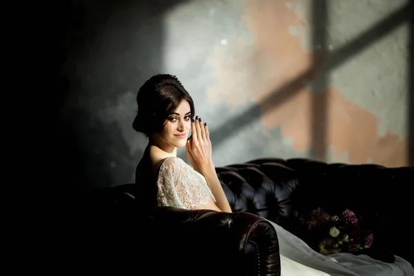 Novia en vestido blanco sentada en la silla en el interior del estudio oscuro como en casa . — Foto de Stock