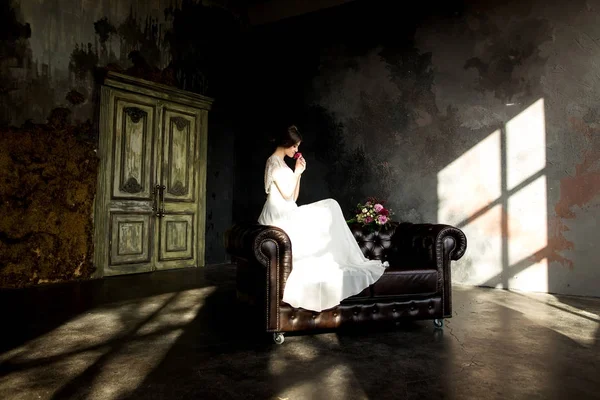 Nevěsta v bílých šatech sedí na židli uvnitř v interiéru tmavě studio jako doma. — Stock fotografie