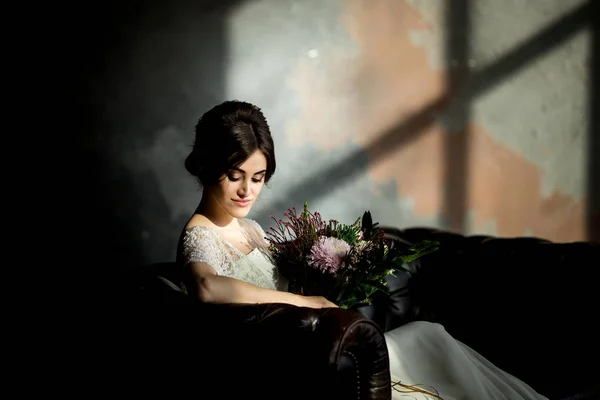 Bride in white dress sitting on chair indoors in dark studio interior like at home. — Stock Photo, Image