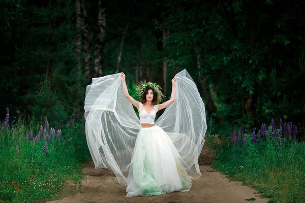 Beautiful bride in white dress and green floral wreath posing in forest, full length