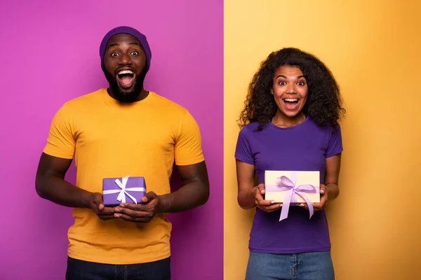 Feliz e alegre casal mostrar o seu presente de Natal. Violeta e amarelo fundo — Fotografia de Stock