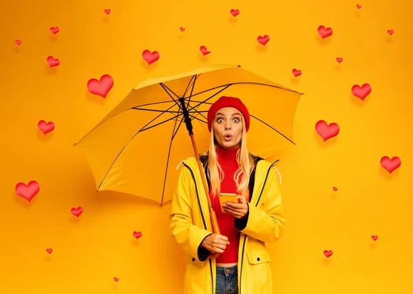 金髪のかわいい女の子は、彼女のスマートフォン上の心の雨のために傘で身を守る。幸せと驚きの表情。背景 — ストック写真
