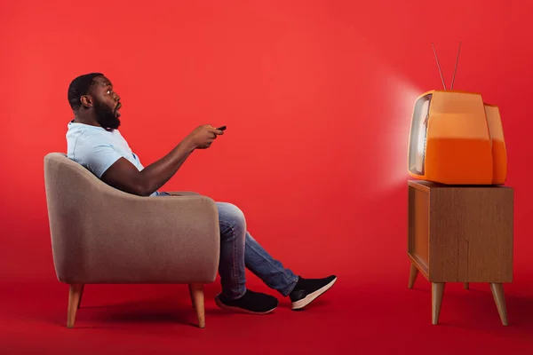 Niesamowity mężczyzna w fotelu ogląda film na zabytkowym telewizorze na czerwonym tle — Zdjęcie stockowe