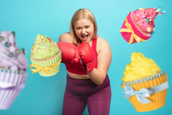 Fat girl in fitness suite does boxing and fight against cupcakes. Cyan background