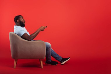 Ev sandalyesindeki televizyon sunucusu kırmızı arkaplanda uzaktan kumandayla kanal değiştirir