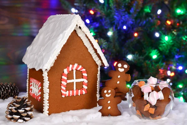 Имбирный домик ручной работы, маленькие человечки и конфеты, новогодняя елка с гирляндой на заднем плане — стоковое фото