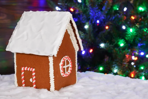 手作りの食べられるジンジャーブレッドハウス、雪の装飾、背景にガーランドと新年の木 — ストック写真