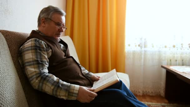 Viejo sentado en el sofá en casa leyendo un libro — Vídeo de stock