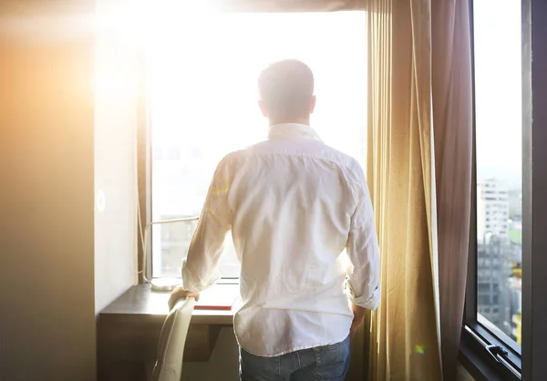 Rear view of young man looking at dawn city scenery in window af — Stock Photo, Image