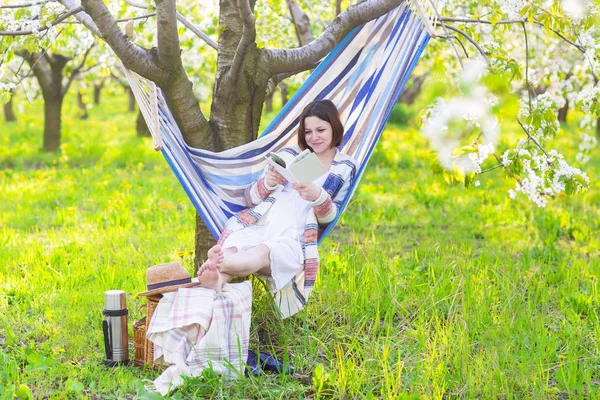 Vacker gravid kvinna sitter i hängmatta i blommande trädgård — Stockfoto