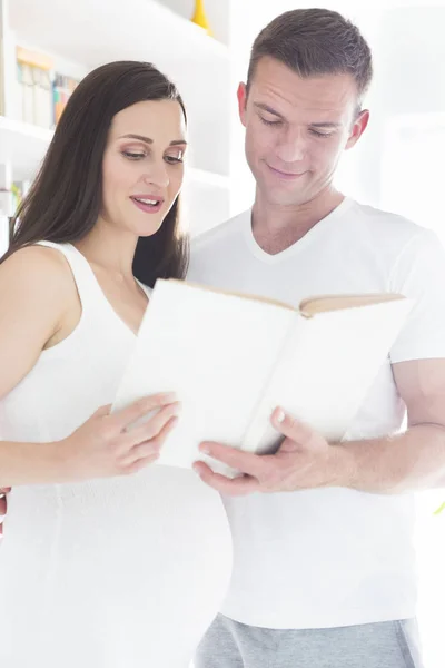 Молодой человек и красивая беременная женщина читают книгу в помещении — стоковое фото