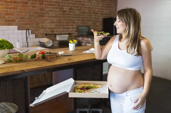 Młoda kobieta w ciąży, jedzenie pizzy w kuchni — Zdjęcie stockowe