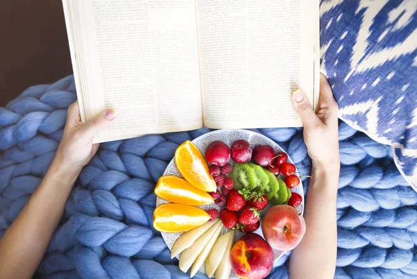 Plate with fresh fruit salad on a blue plaid and book. Woman rea — Stock Photo, Image