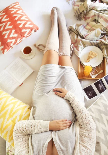 Mujer embarazada con libro, té, pastel relajante en casa — Foto de Stock