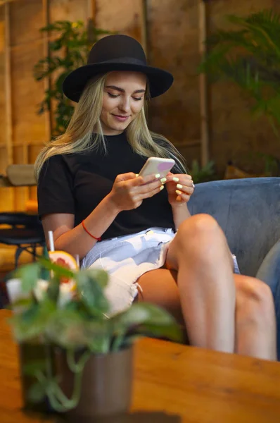 Menina loira em saia jeans branco, blusa e chapéu preto, sentado a — Fotografia de Stock