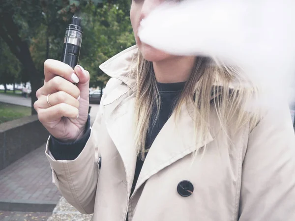 Blond girl in beige trench coat vaping fruit e-liquid with very