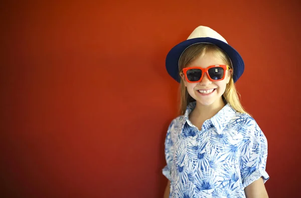 Niño bastante emotional usar un sombrero y gafas de sol en un respaldo rojo — Foto de Stock