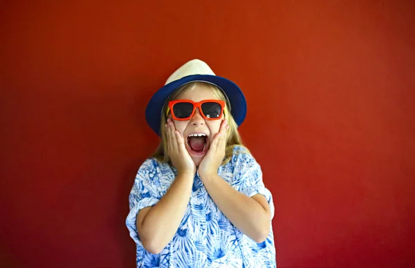 Niño bastante emotional usar un sombrero y gafas de sol en un respaldo rojo — Foto de Stock