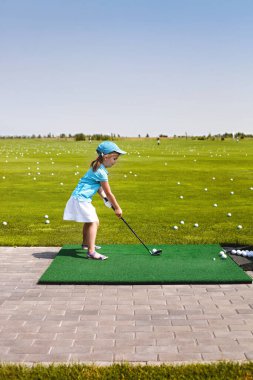 Güneşli bir günde yeşil sahada golf antrenmanında küçük bir kızın topa vurmaya hazırlanışının yan görüntüsü.