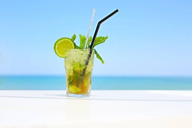 Beyaz masadaki renkli yeşil kokteyl kireç dilimi ile süslenmiş şık cam ve mavi arka planda izole siyah tüp ile taze nane.