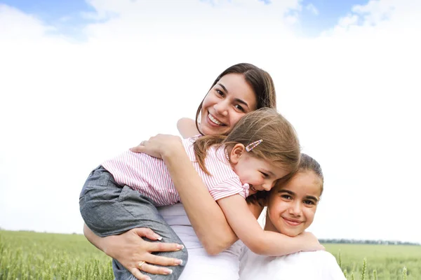 快乐的年轻母亲笑着看着摄像机 背对着绿色的田野和天空拥抱着小女儿 — 图库照片