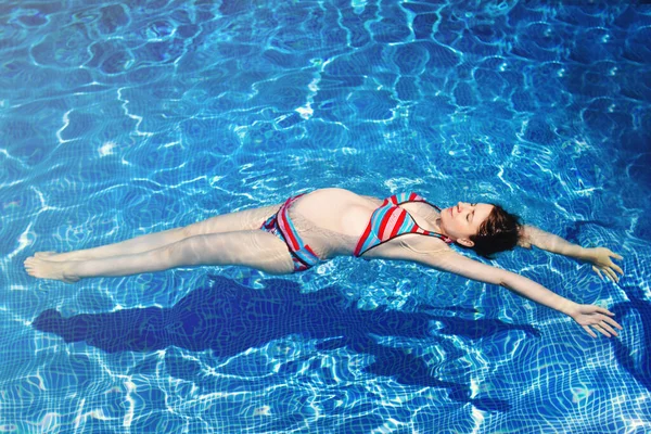 从上面看 穿着条纹比基尼的轻松自在的孕妇漂浮在度假胜地游泳池清澈的水中 — 图库照片