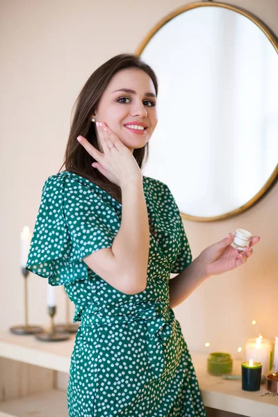 Happy brunette in green polka dot dress moisturizing face skin with cream looking at camera