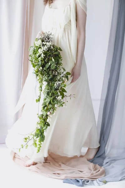 穿着雅致的白色婚纱的光着脚的女人 她站在透明的彩色窗帘中间 一束束着白色的小花和绿色的叶子 — 图库照片