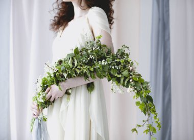 Çıplak ayaklı, zarif, beyaz gelinlikli, büyük bir buket beyaz çiçekli, yeşil yapraklı, şeffaf pastel perdelerin arasında duran bir kadın.